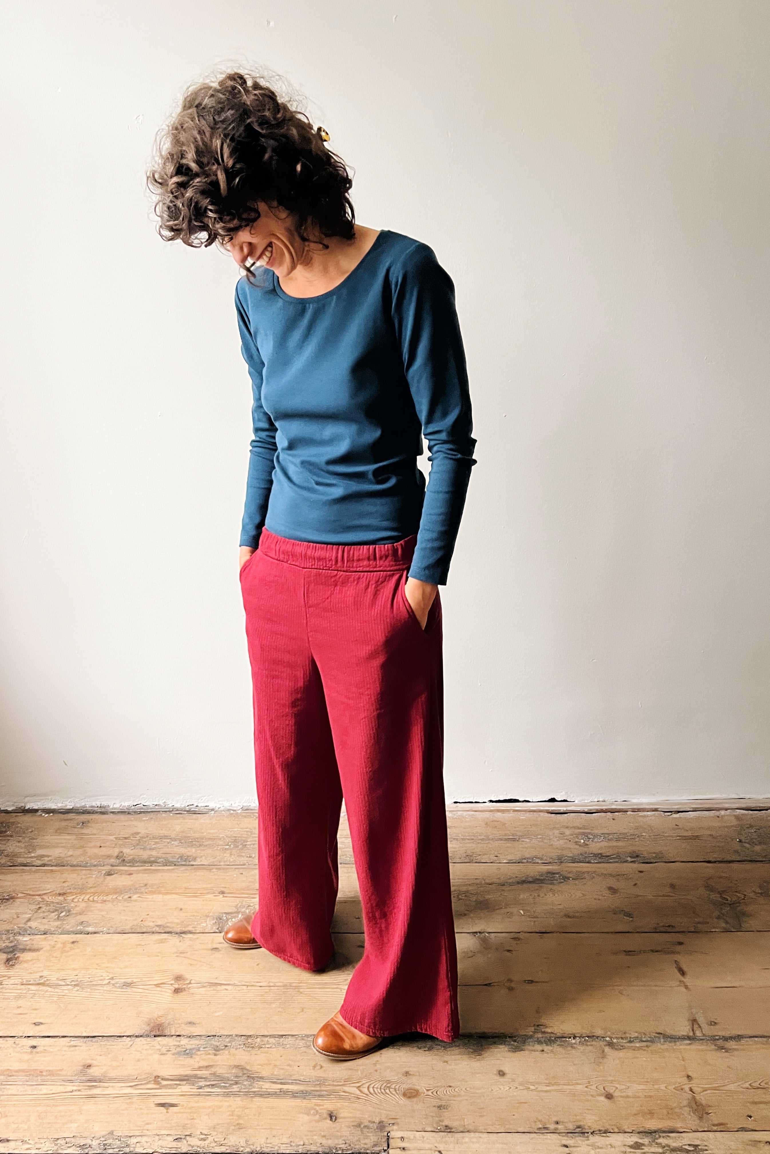 Person wearing an ocean blue longsleeve top and red pants standing on a wooden floor against a white wall.