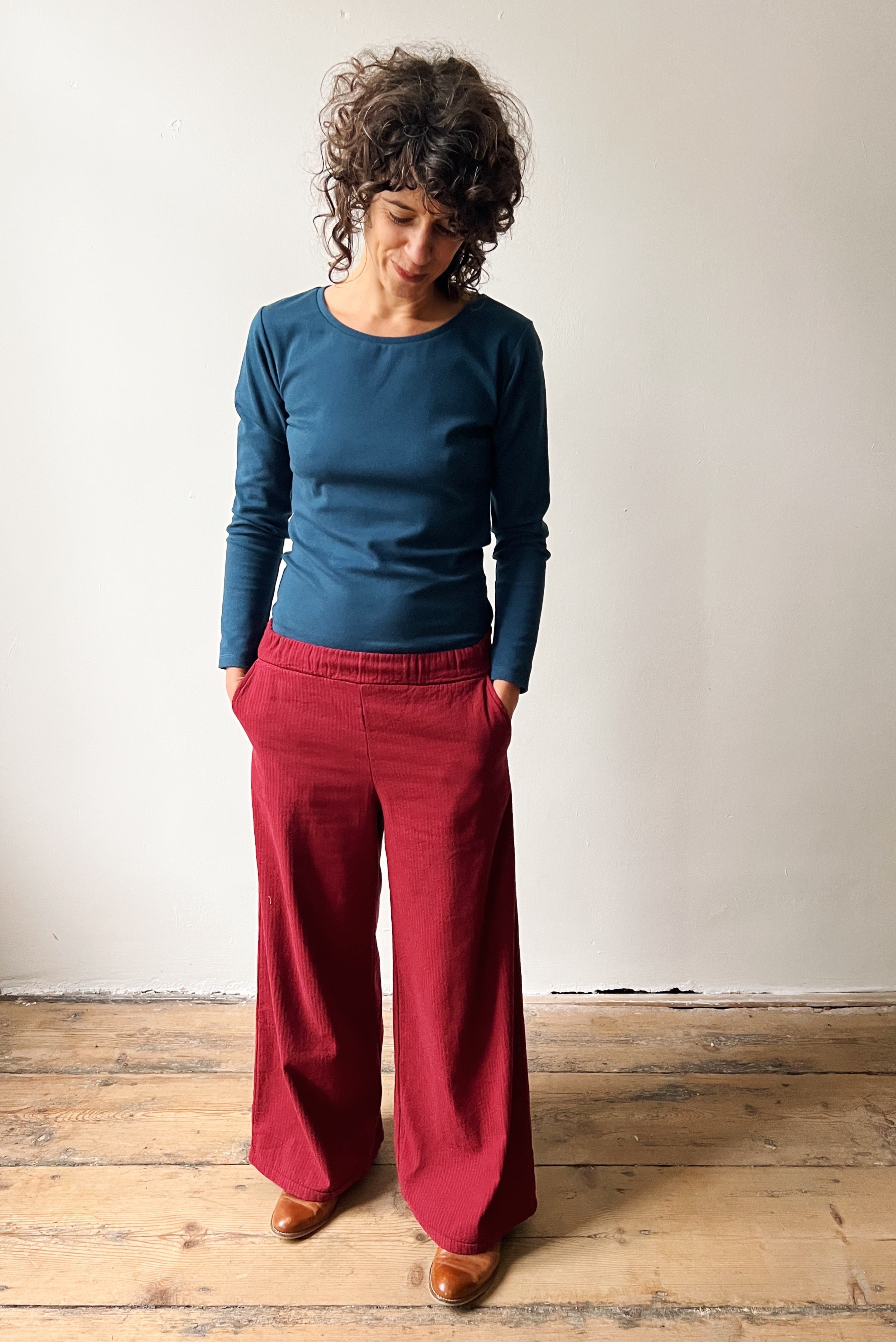 Person wearing an ocean blue longsleeve top and wide red pants standing on a wooden floor against a white wall.