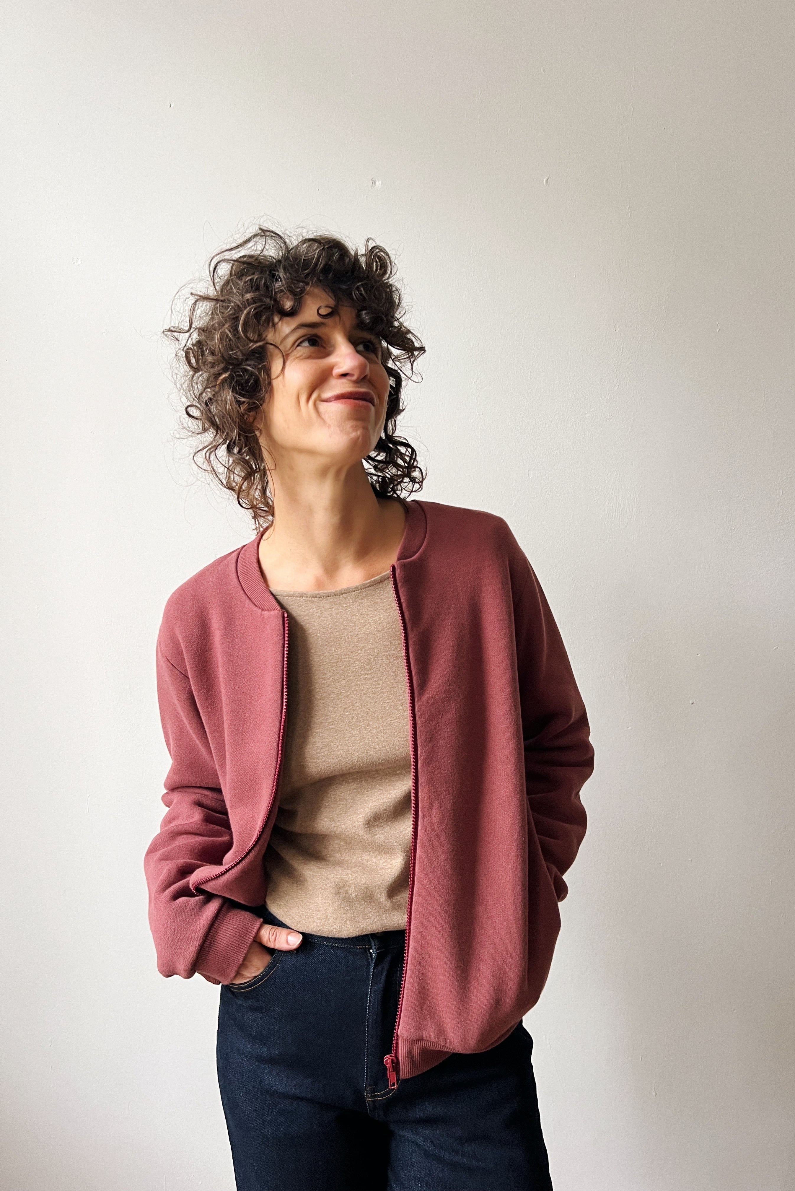 Woman wearing a figue bomber over a beige top and blue jeans against a plain background