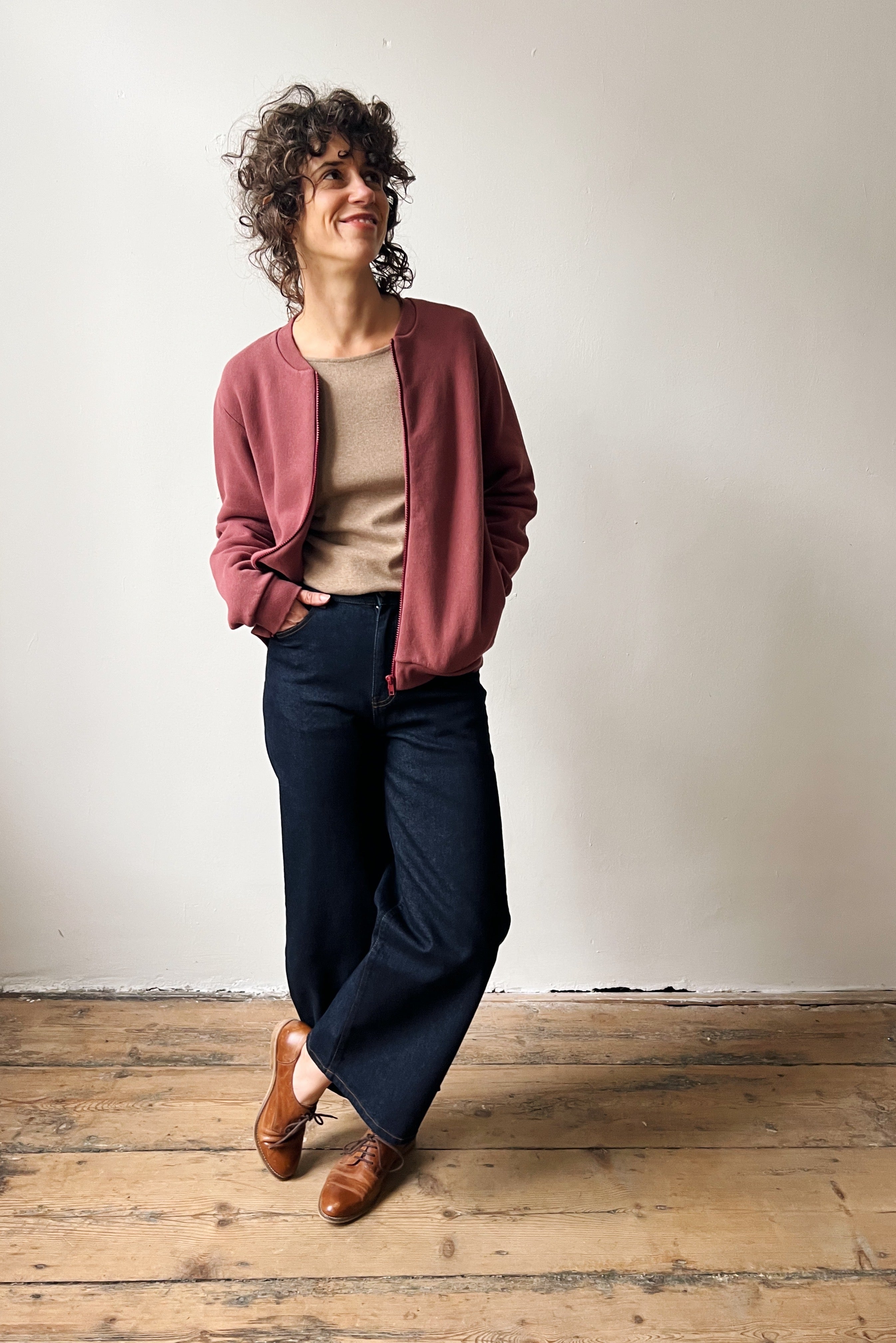 Woman wearing a figue bomber, beige top, and dark pants on a wooden floor.