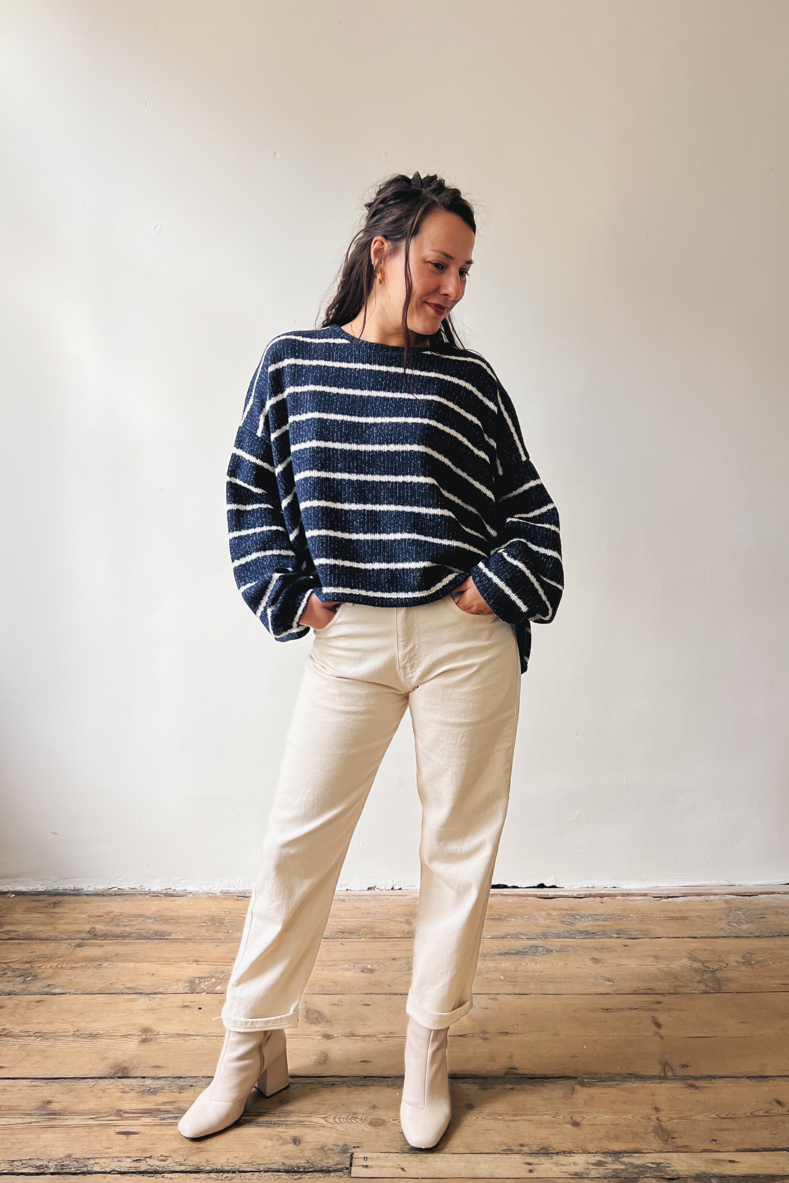 Woman wearing a blue and white striped sweater with oat jeans and white boots against a plain wall.