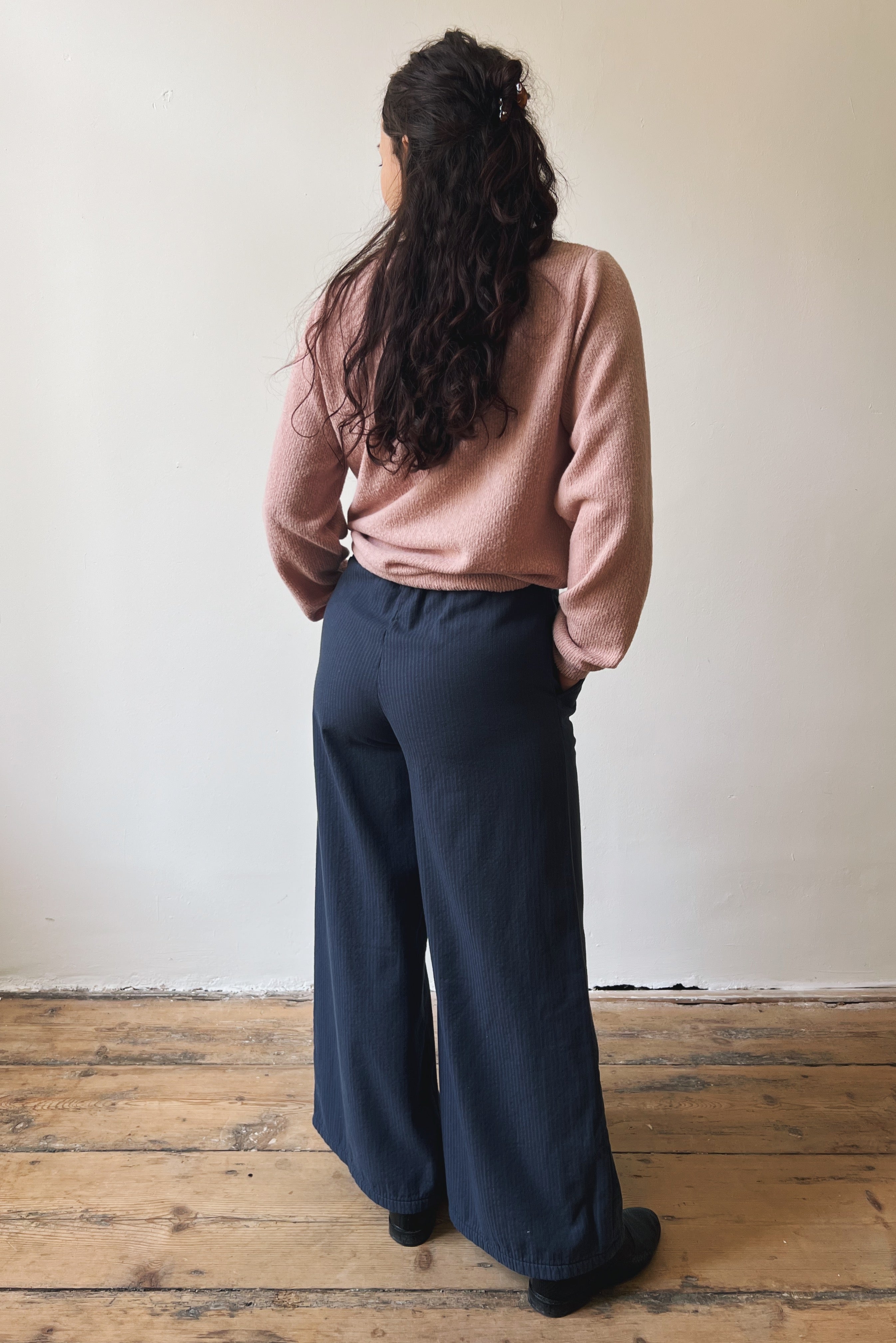 Woman wearing a pink shirt and dark blue wide-leg pants standing against a plain wall.