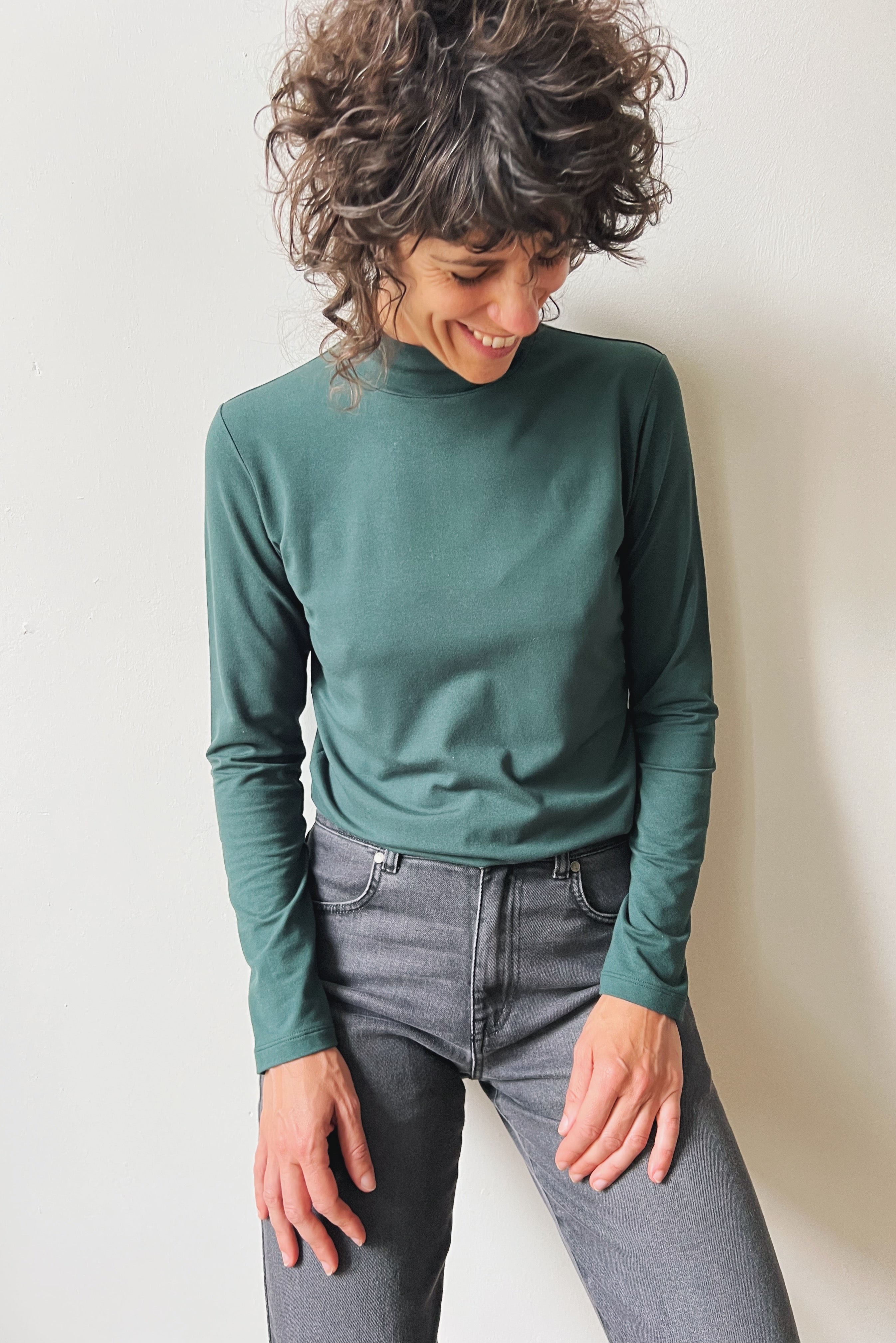 Woman wearing a pine turtleneck shirt and gray jeans against a plain background
