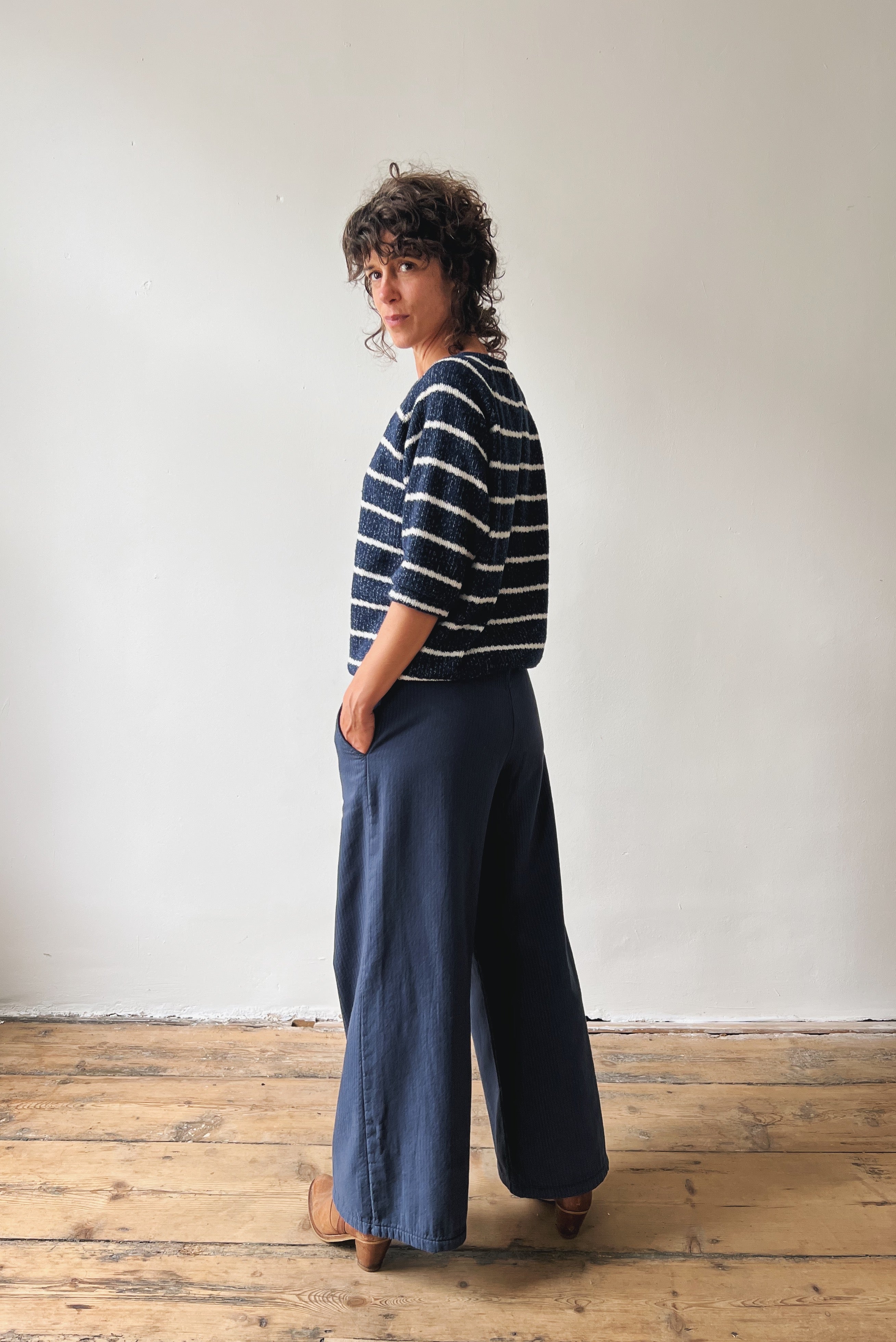 Woman wearing a striped sweater and wide-leg blue pants standing against a white wall.