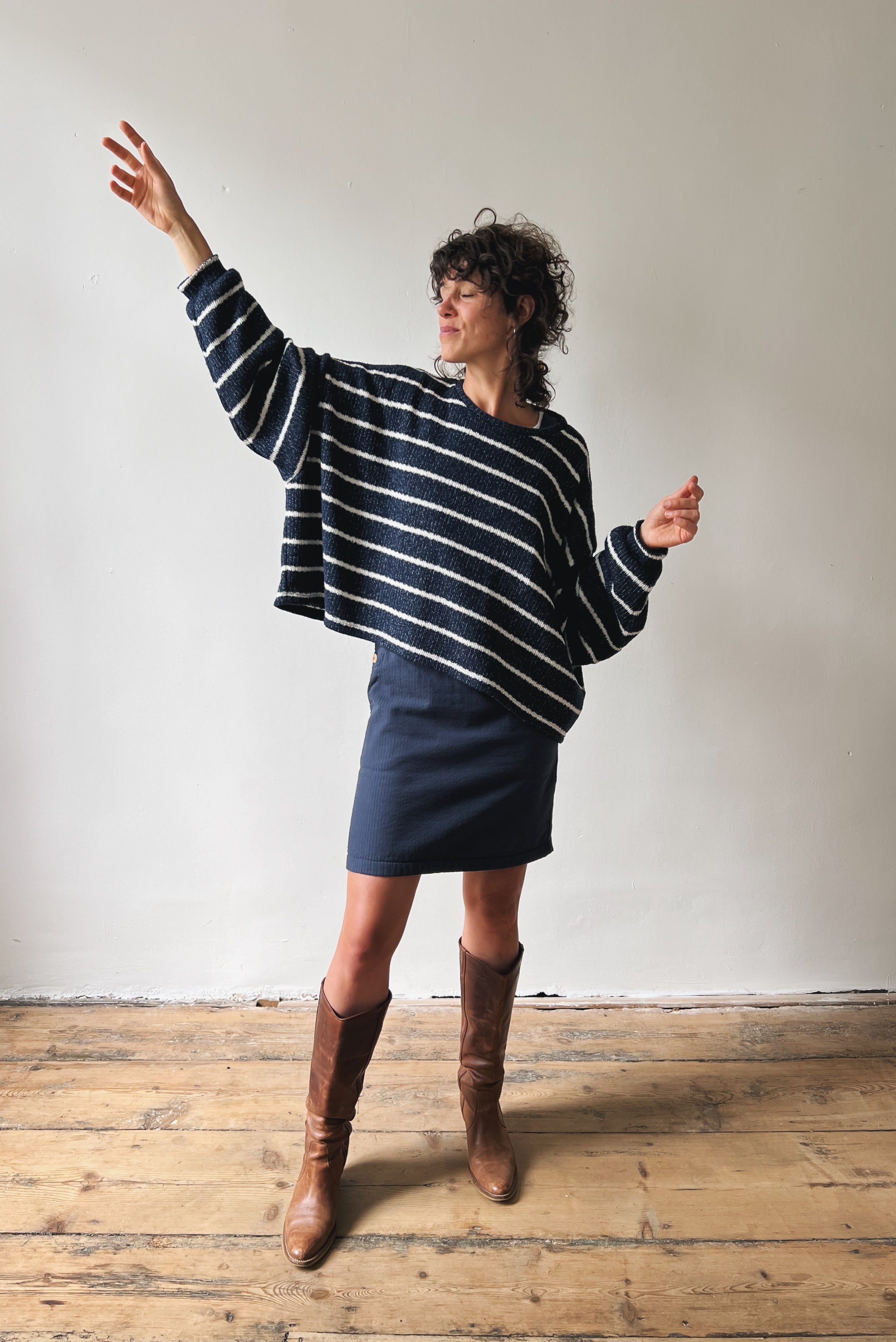 Person wearing a blue and white striped sweater and blue skirt with brown knee-high boots on a wooden floor.