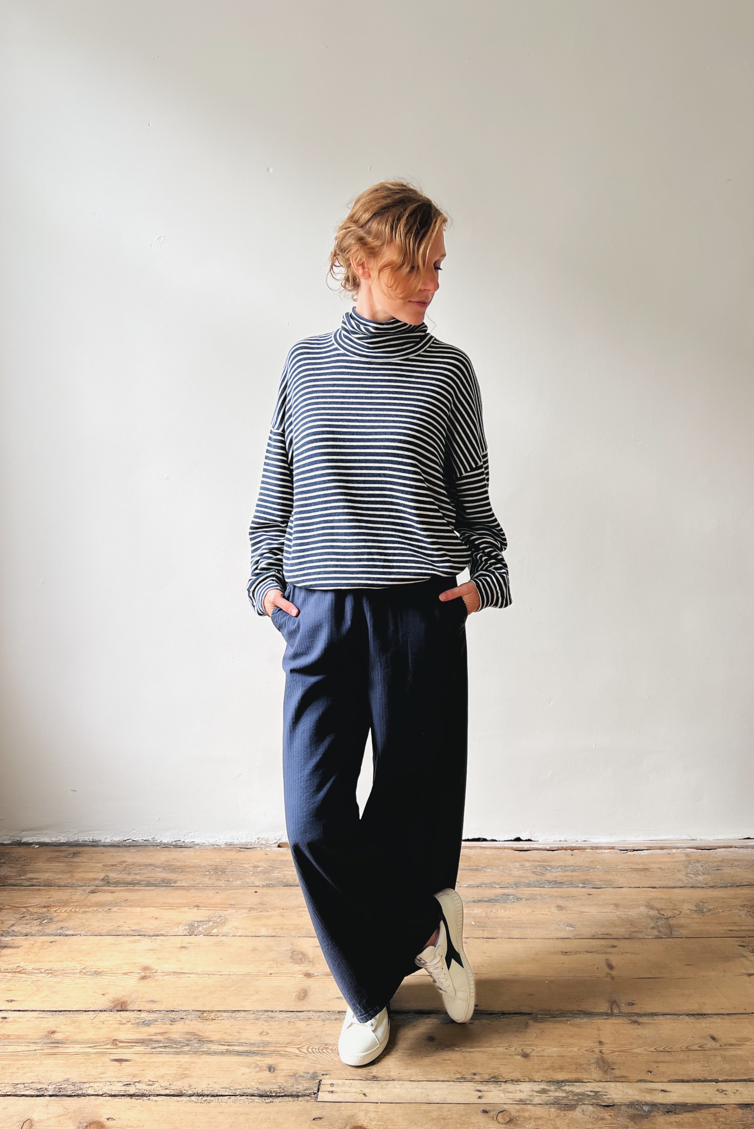 Person wearing a striped sweater and dark blue pants standing on a wooden floor with a white wall background
