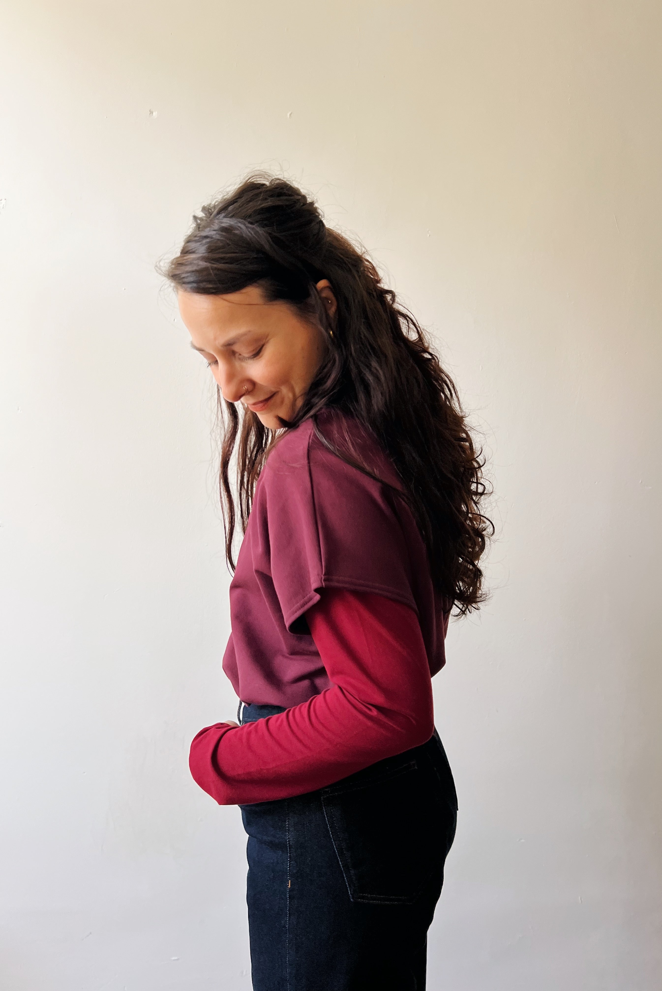 Woman wearing an aubergine sweater, red longsleeve top and dark jeans against a plain background