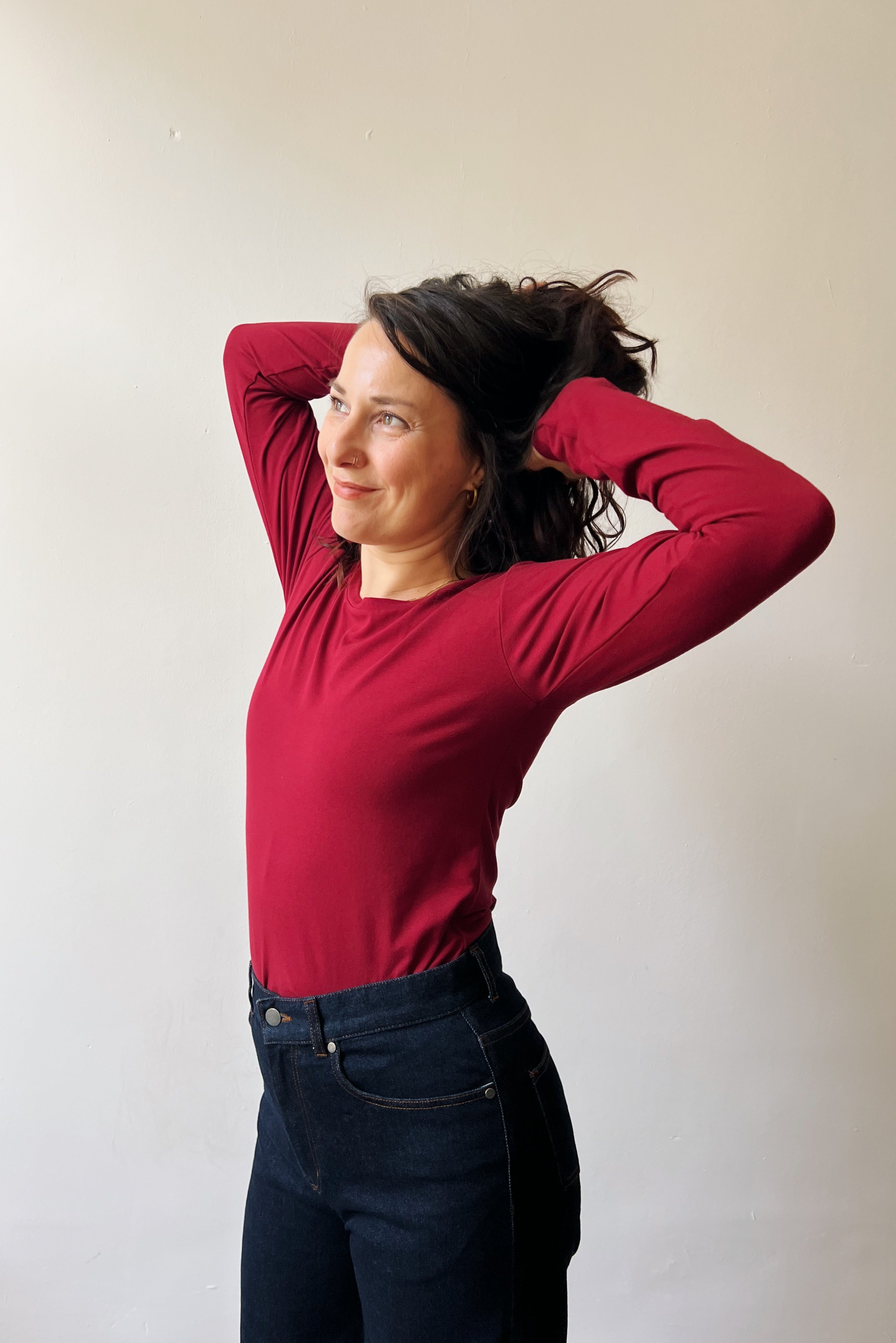 Woman wearing a red long-sleeve top and dark jeans against a plain background