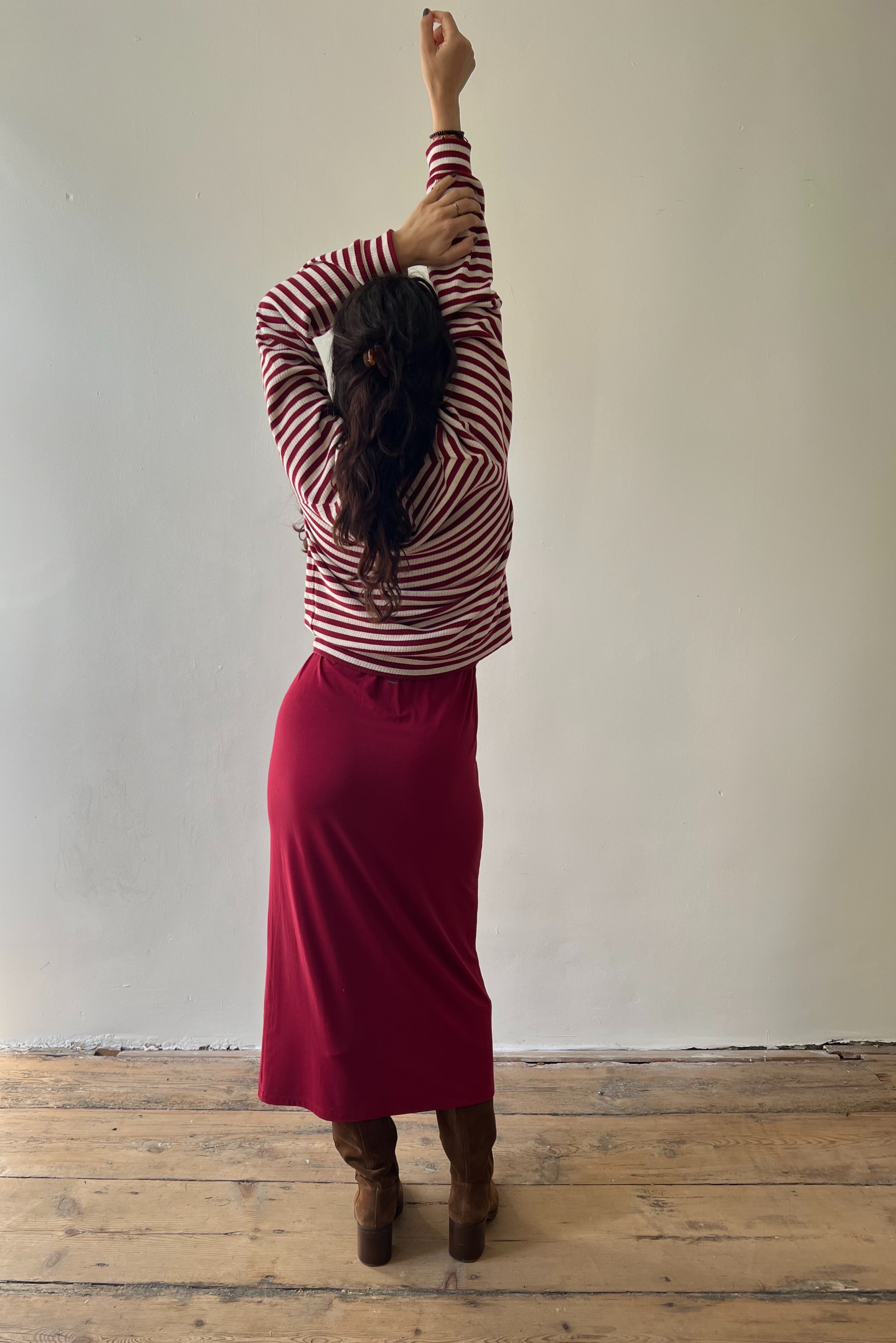 Person wearing a striped shirt and red skirt stretching against a plain wall.