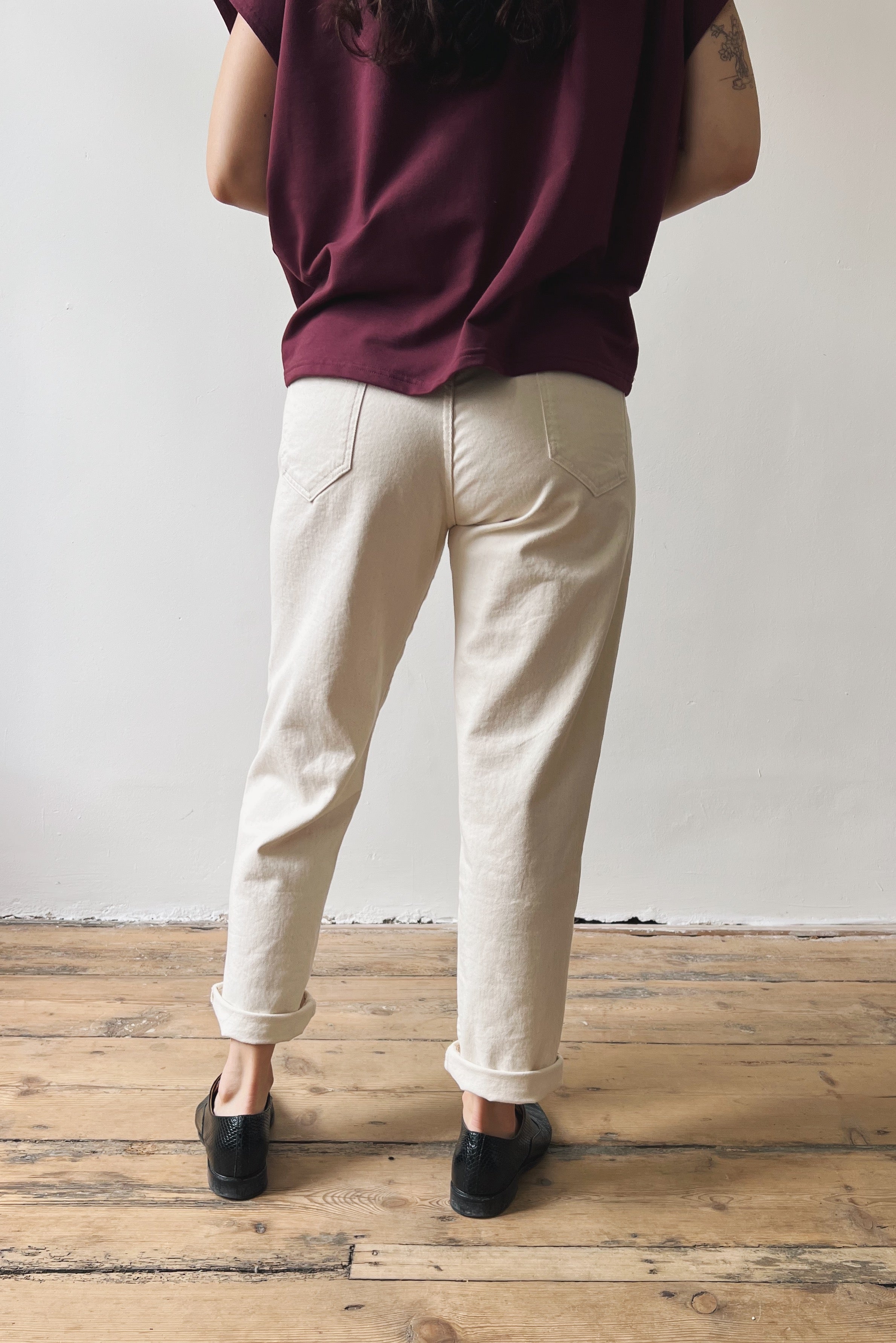 Person wearing an aubergine top and oat jeans standing on a wooden floor with a white wall background