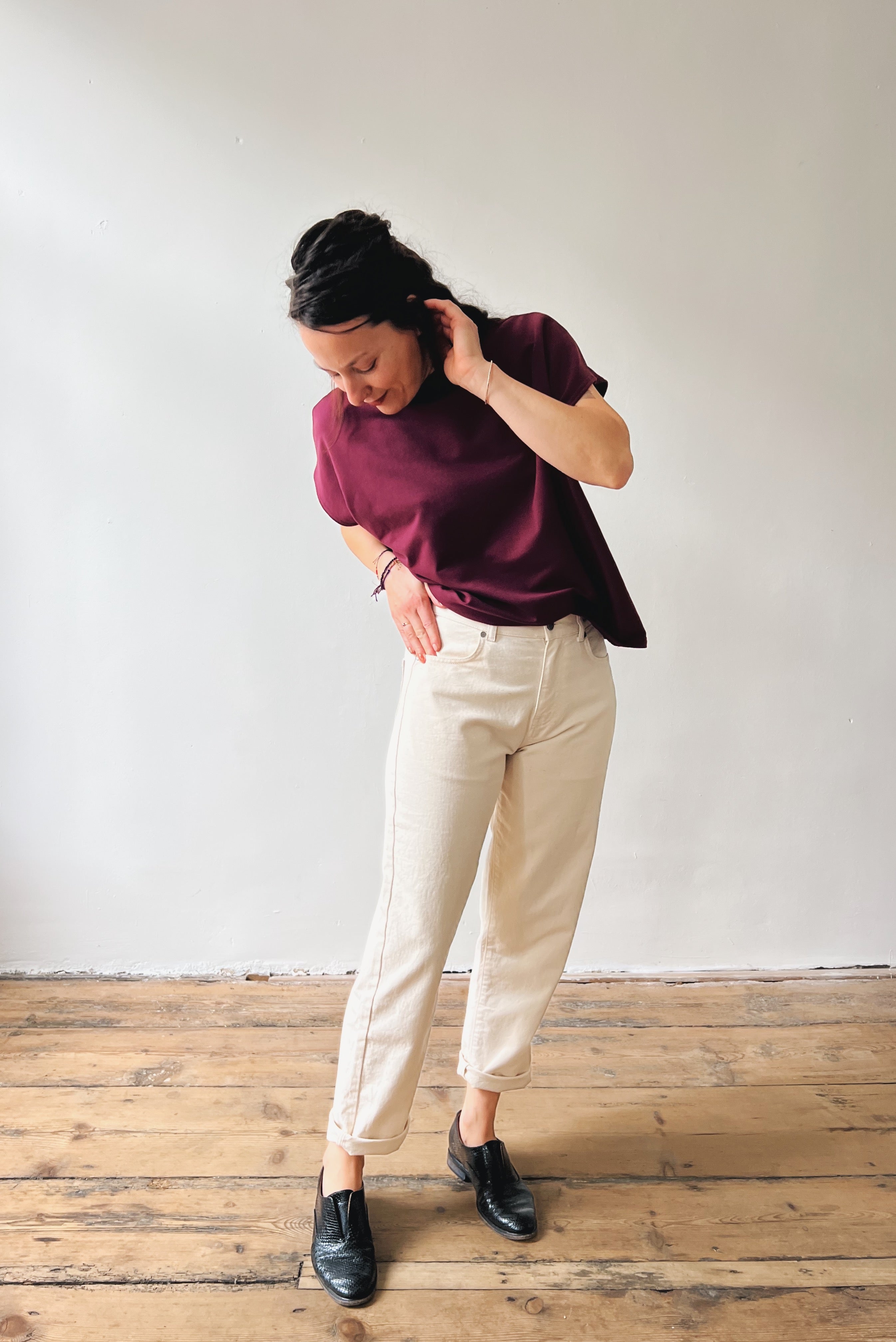 Person wearing a aubergine shirt and oat jeans standing on a wooden floor with a white wall background