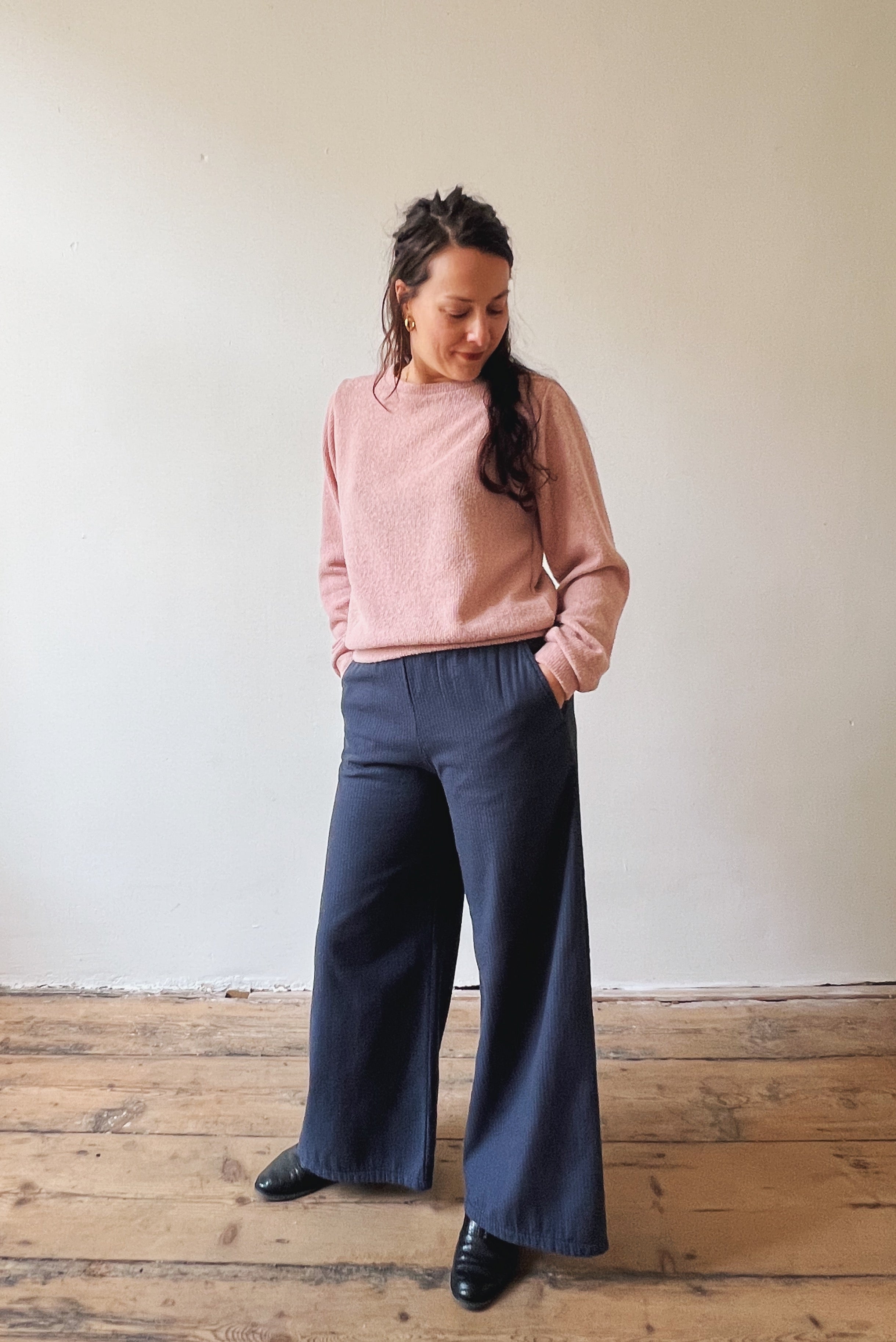 Person wearing a pink sweater and blue pants standing against a plain wall.