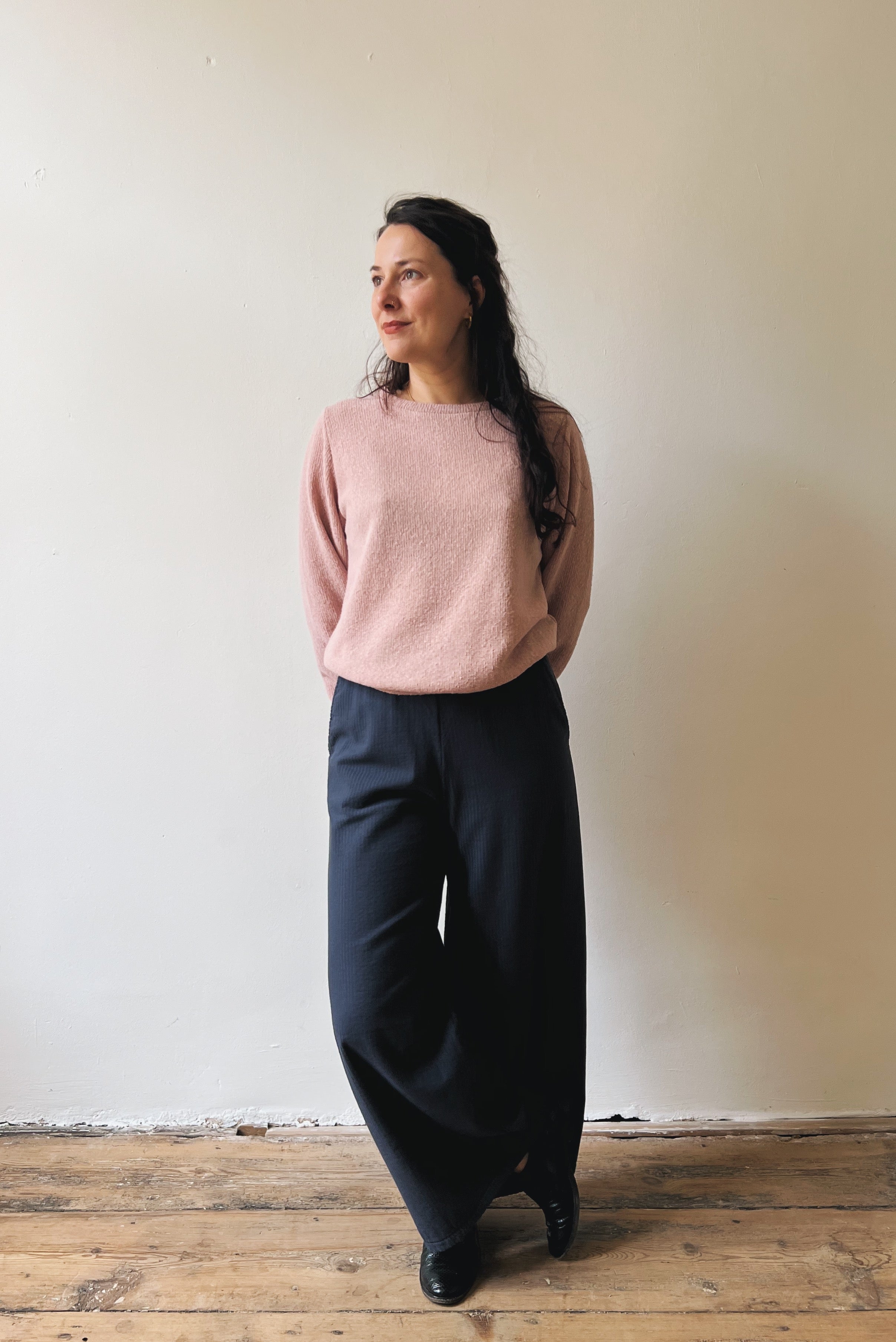Woman wearing a pink sweater and dark pants standing against a plain wall.