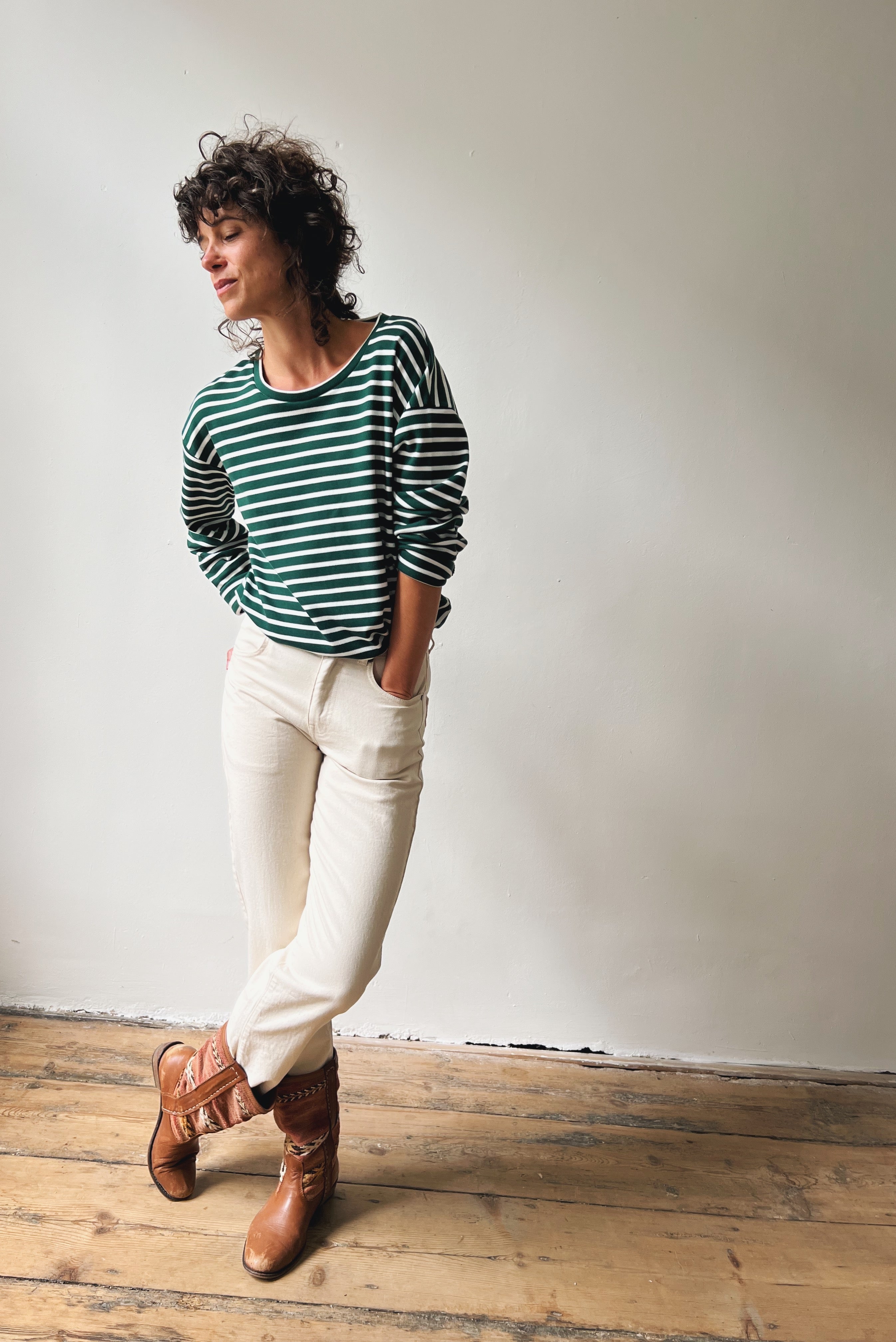 Person wearing a green and black striped sweater with oat jeans and brown shoes against a plain wall.