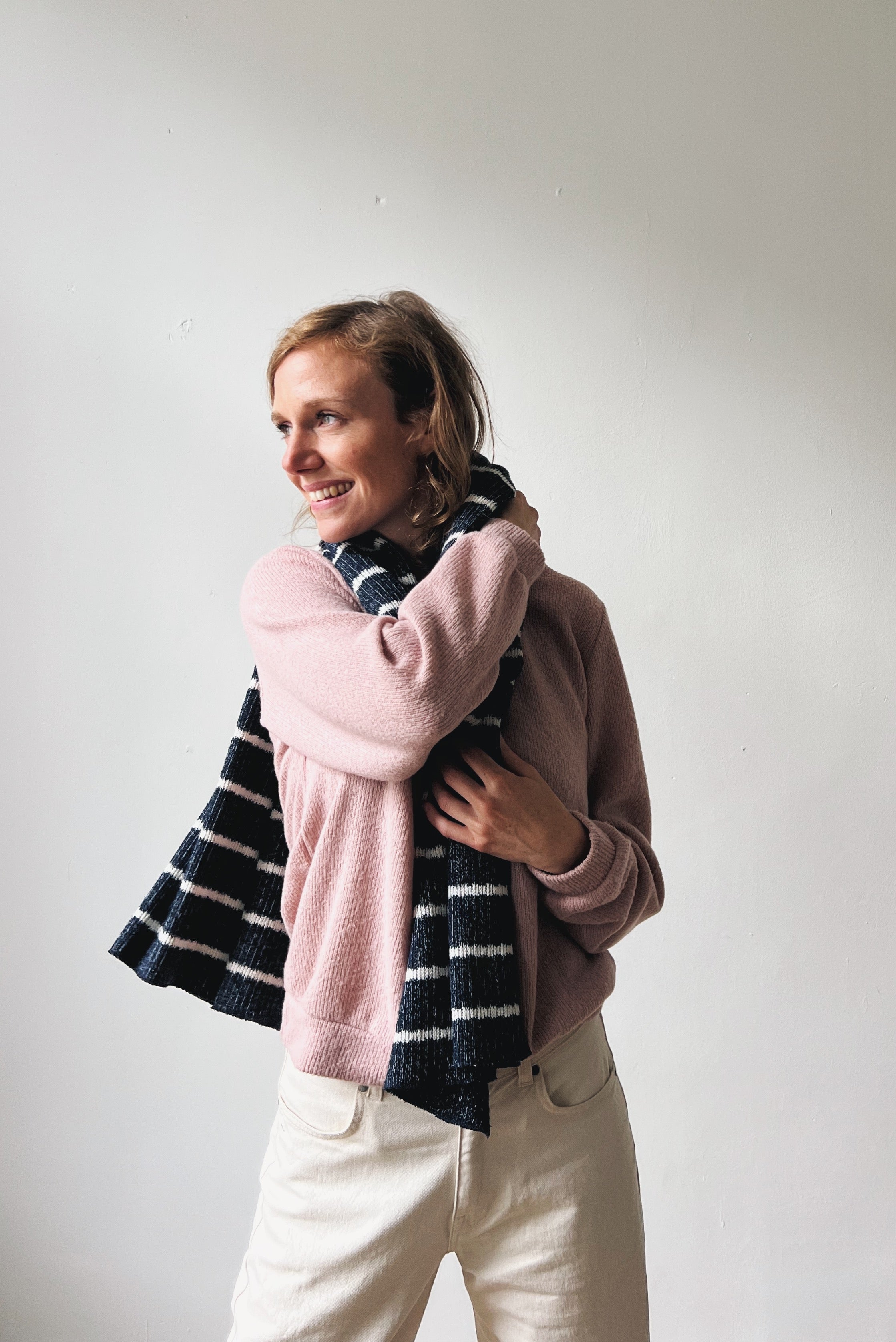 Woman wearing a pink sweater and oat jeans with a blue-white striped scarf against a plain background