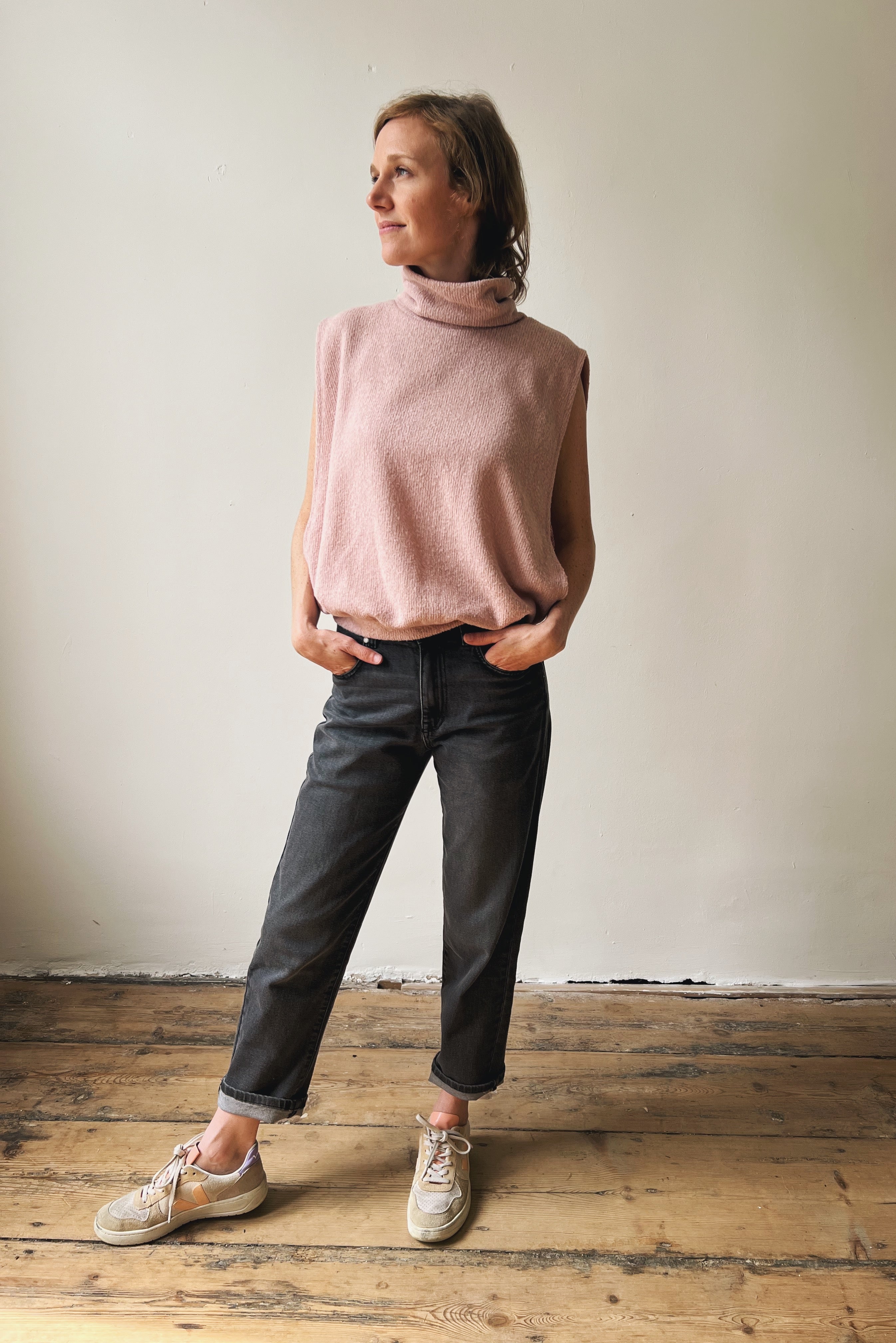 Woman wearing a pink sleeveless sweater and dark grey jeans standing on a wooden floor.
