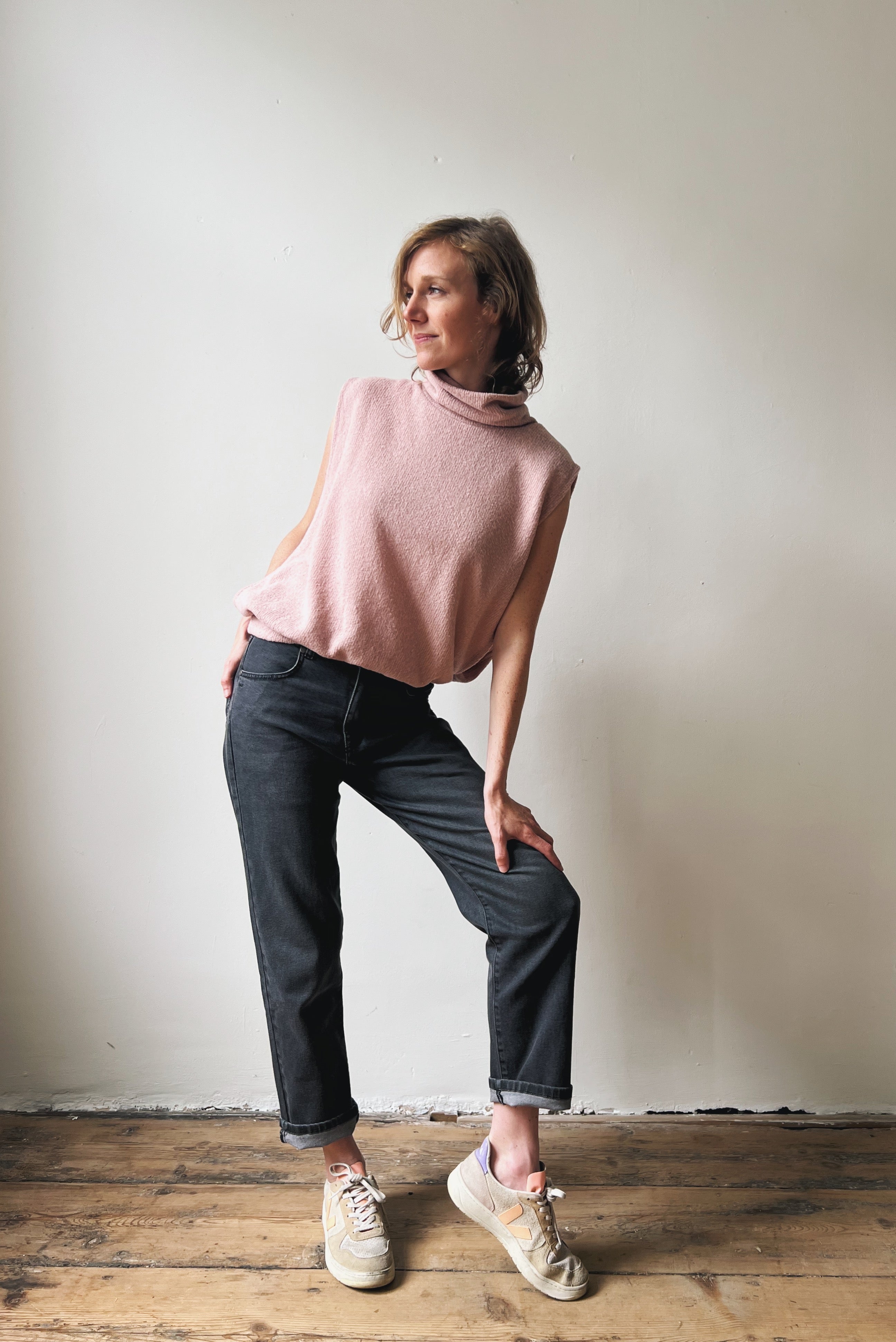 Woman wearing a pink sleeveless top and dark grey jeans standing against a plain wall.