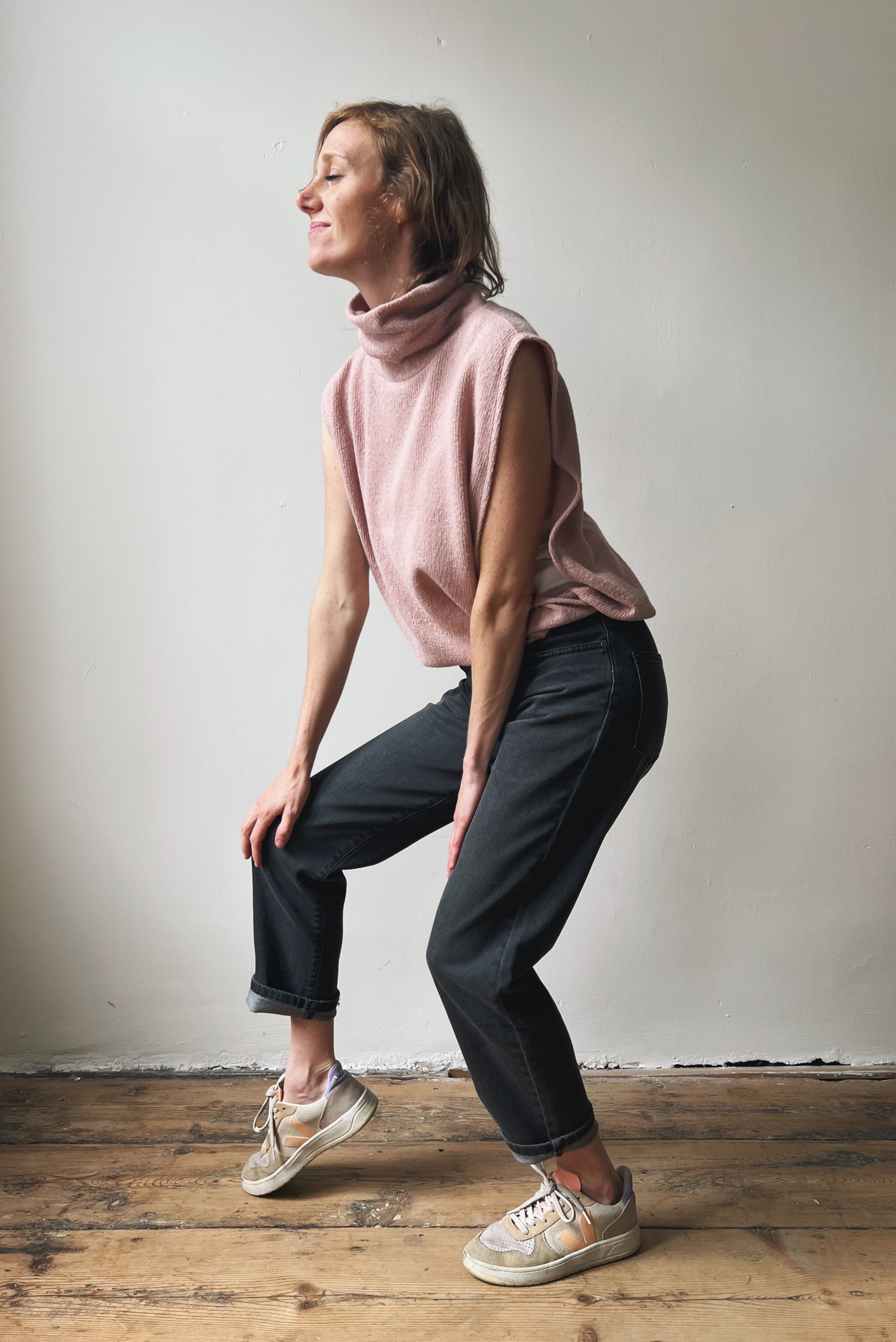 Woman in a pink sleeveless sweater and grey jeans standing on a wooden floor against a plain wall.