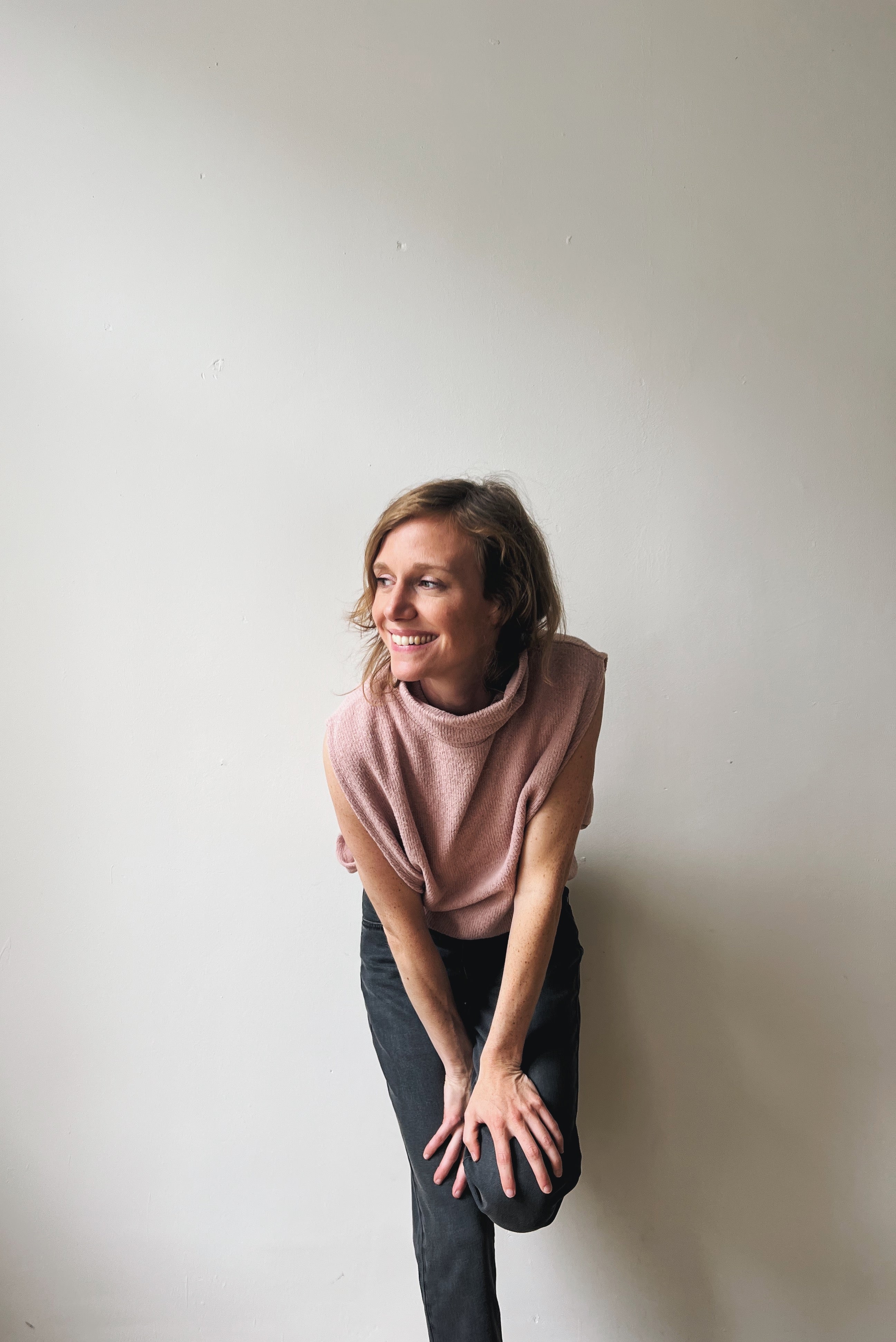 Woman in a pink sleeveless sweater and grey jeans leaning against a plain wall