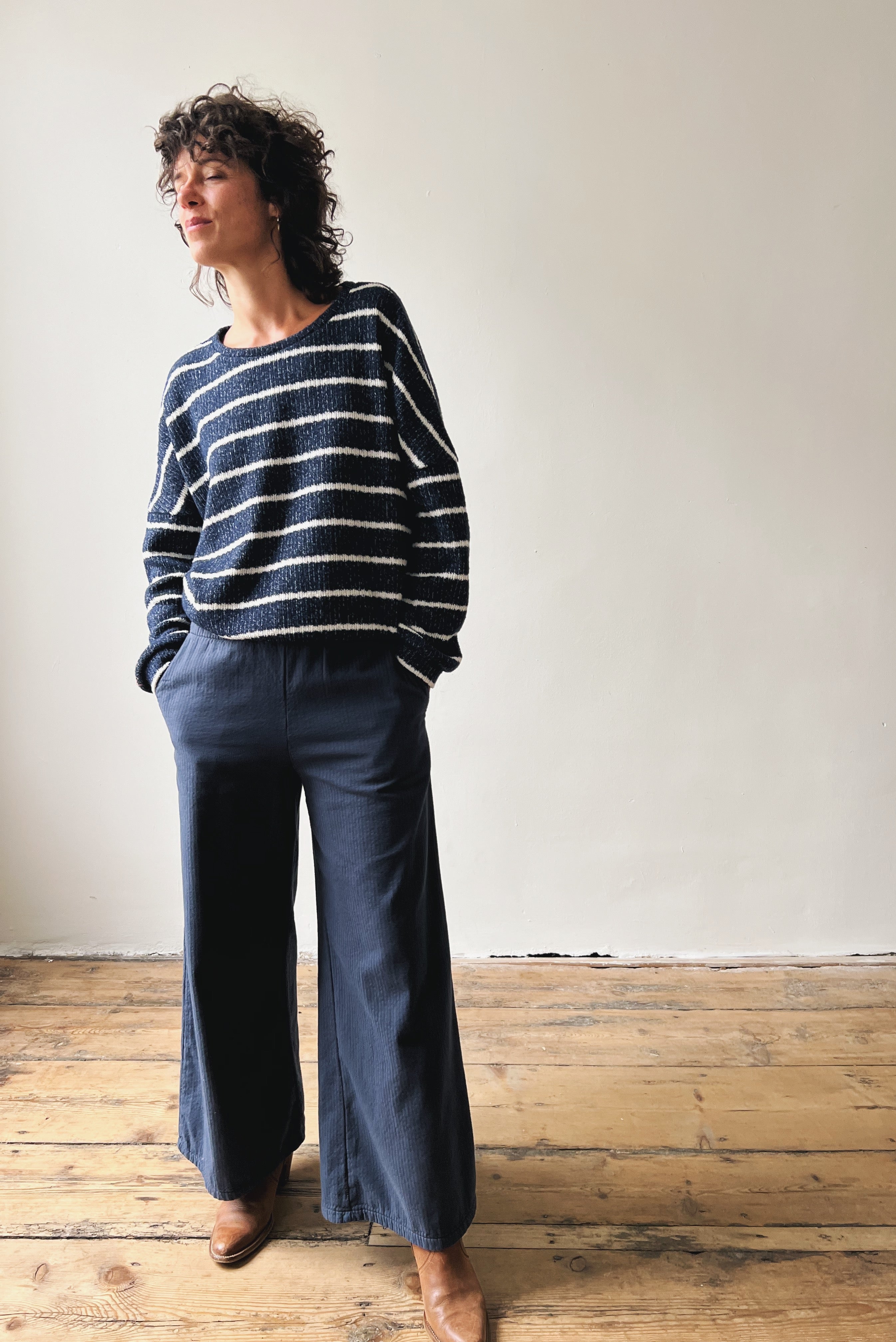 Woman wearing a striped sweater and dark blue pants standing against a plain wall.