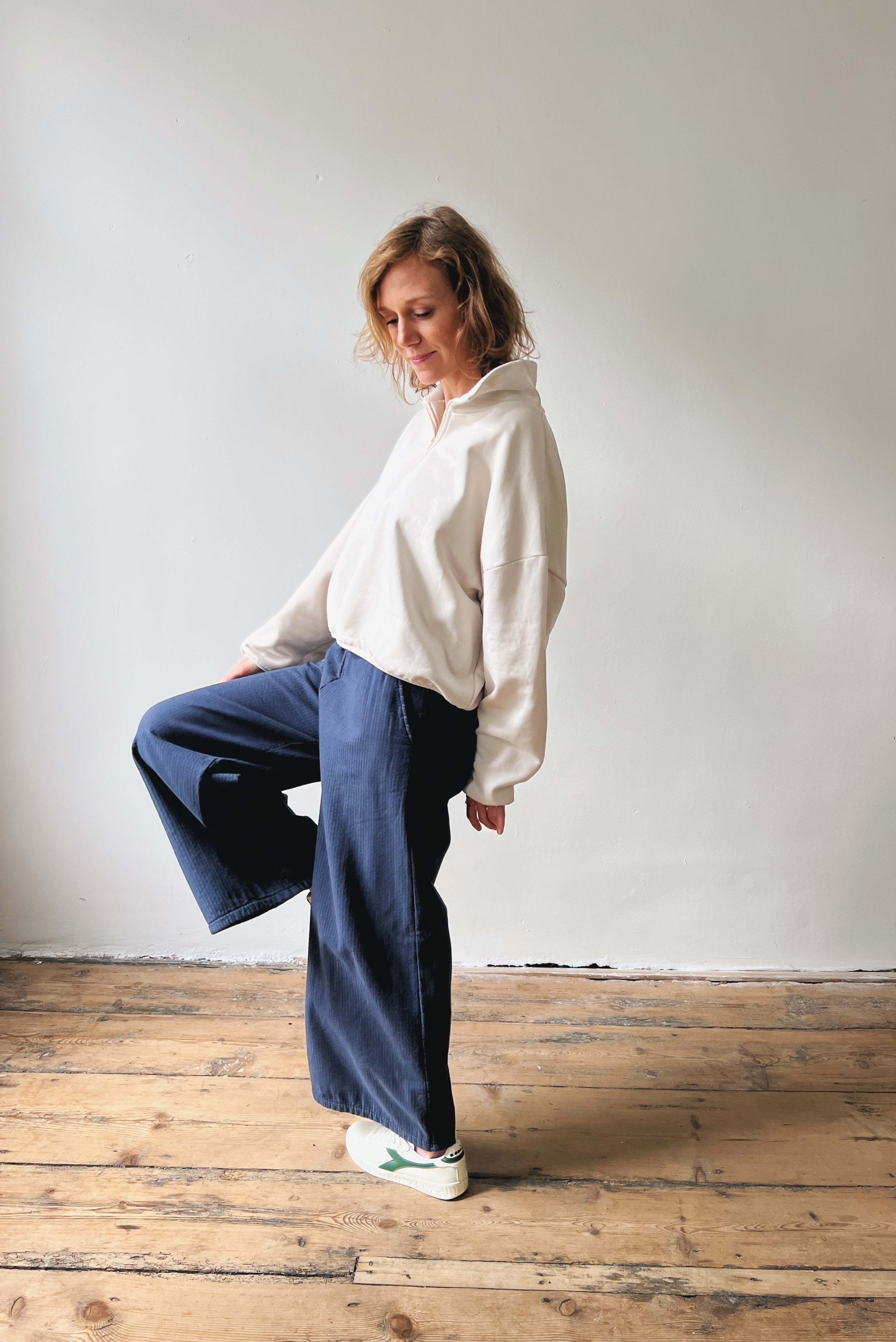 Person wearing a white sweater and wide-leg blue pants standing on a wooden floor against a plain wall.