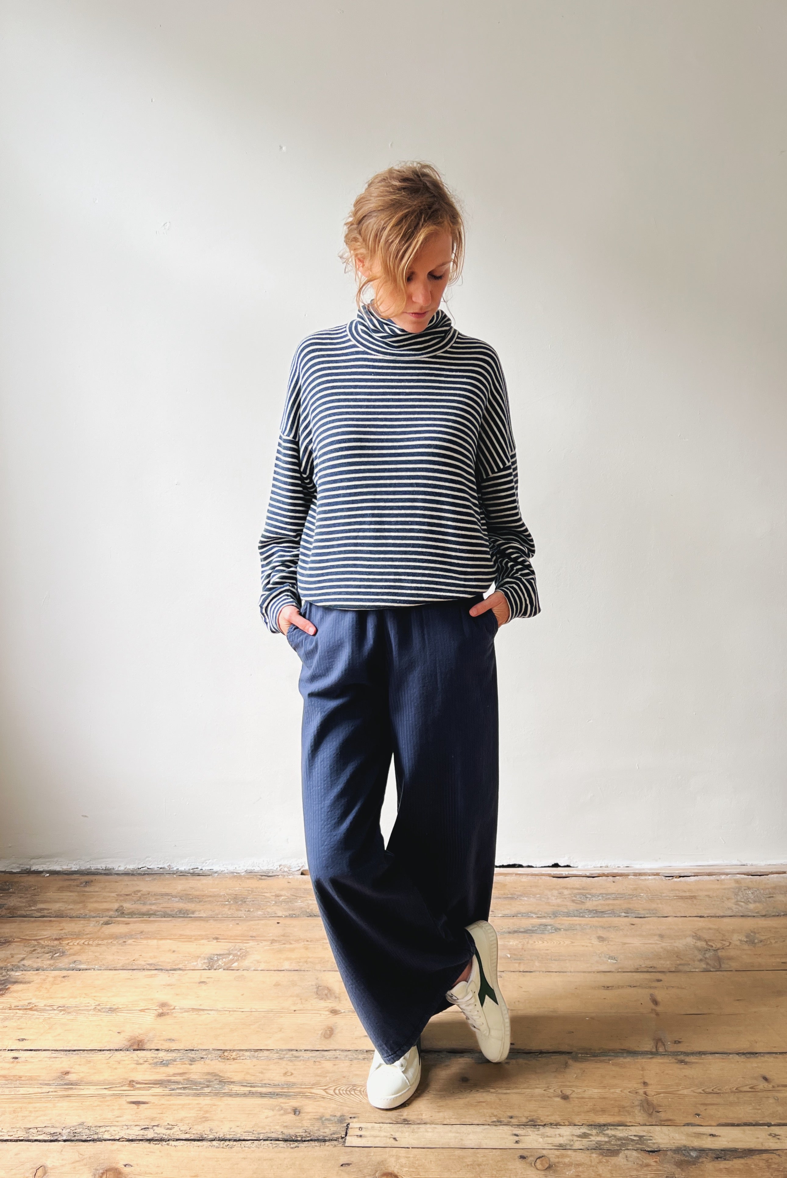 Person wearing a striped sweater and navy pants standing on a wooden floor with a white wall background