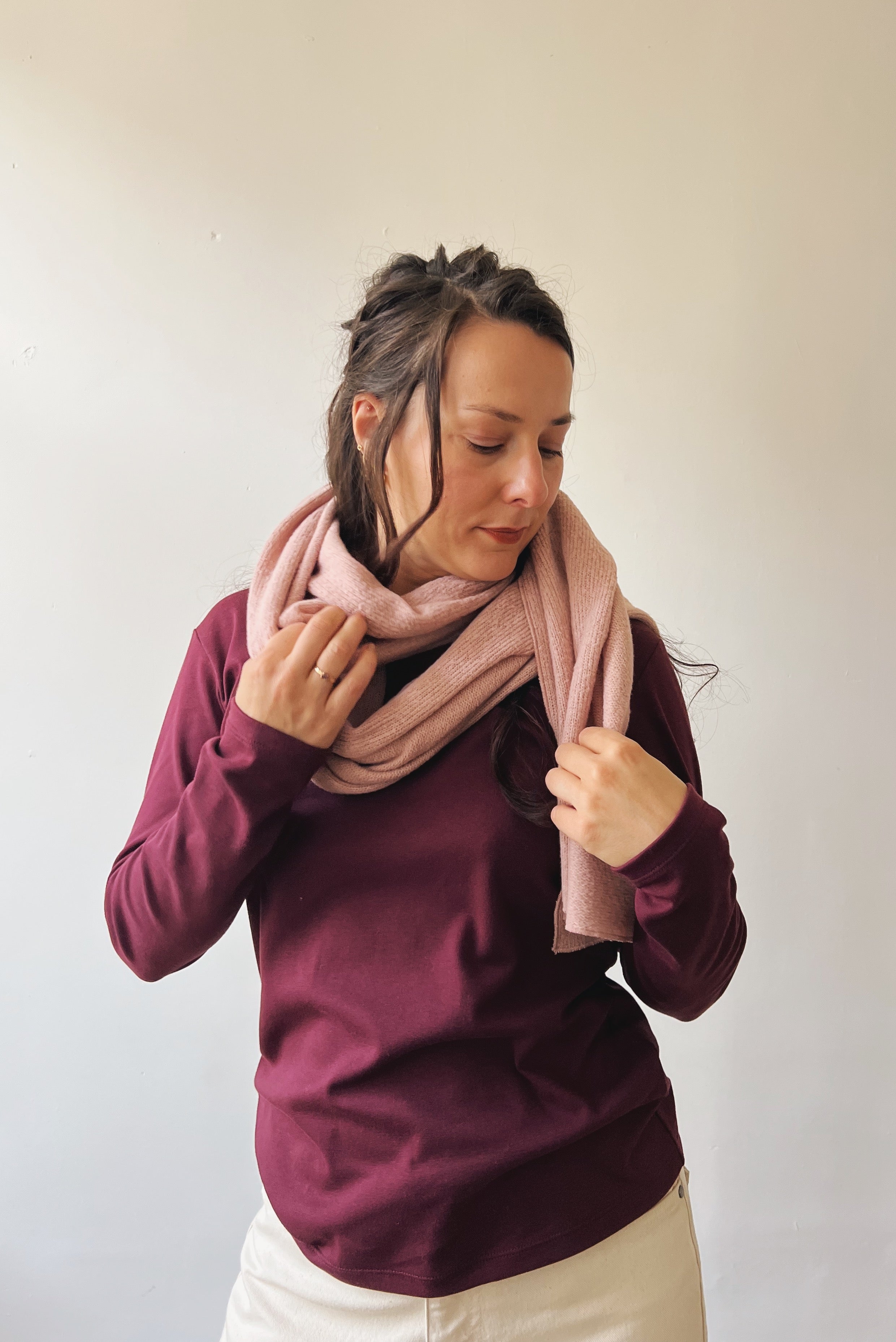 Woman wearing a pink scarf and aubergine longsleeve against a plain background