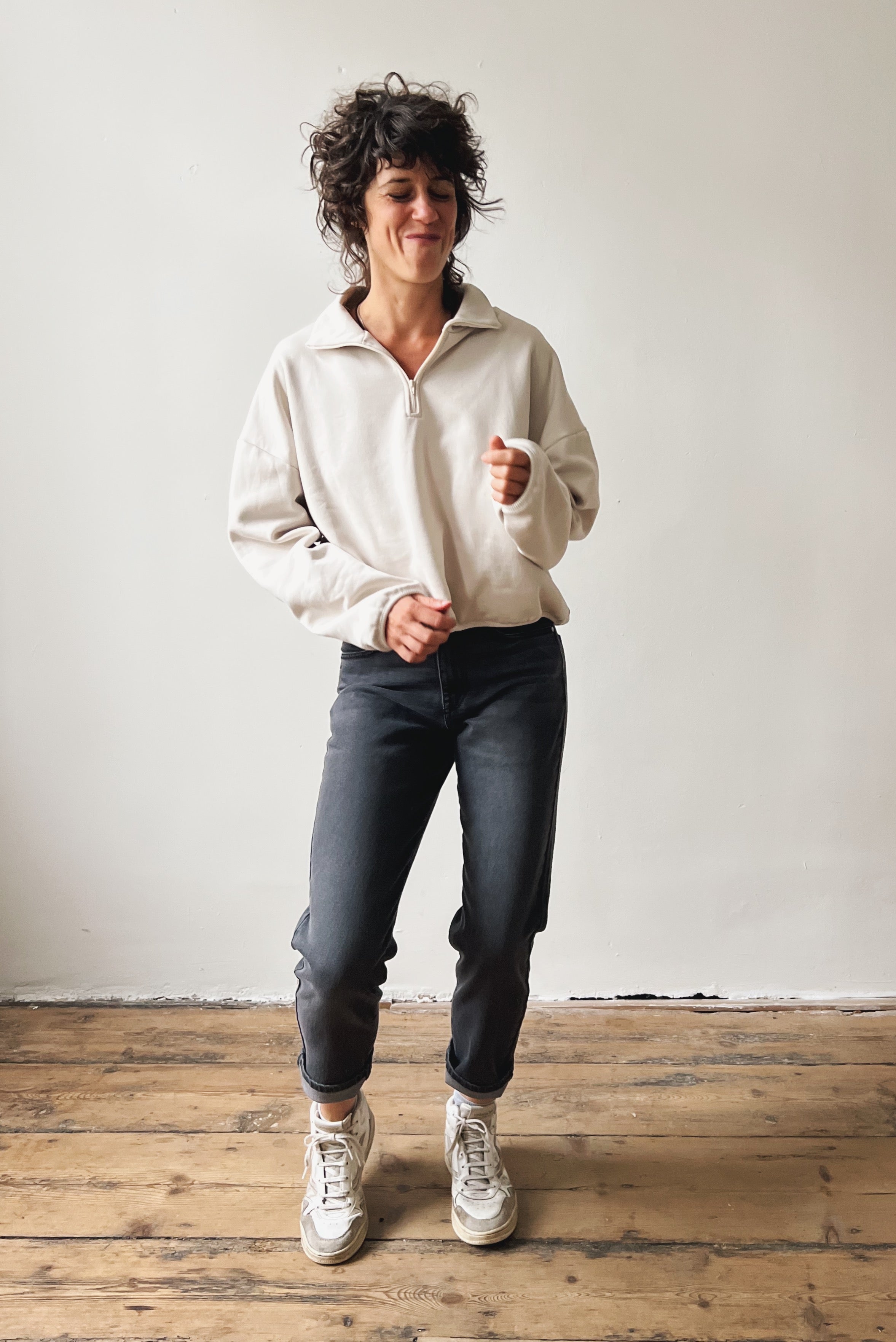 Woman wearing a beige sweater and dark grey boyfriend jeans standing on a wooden floor with a white wall background