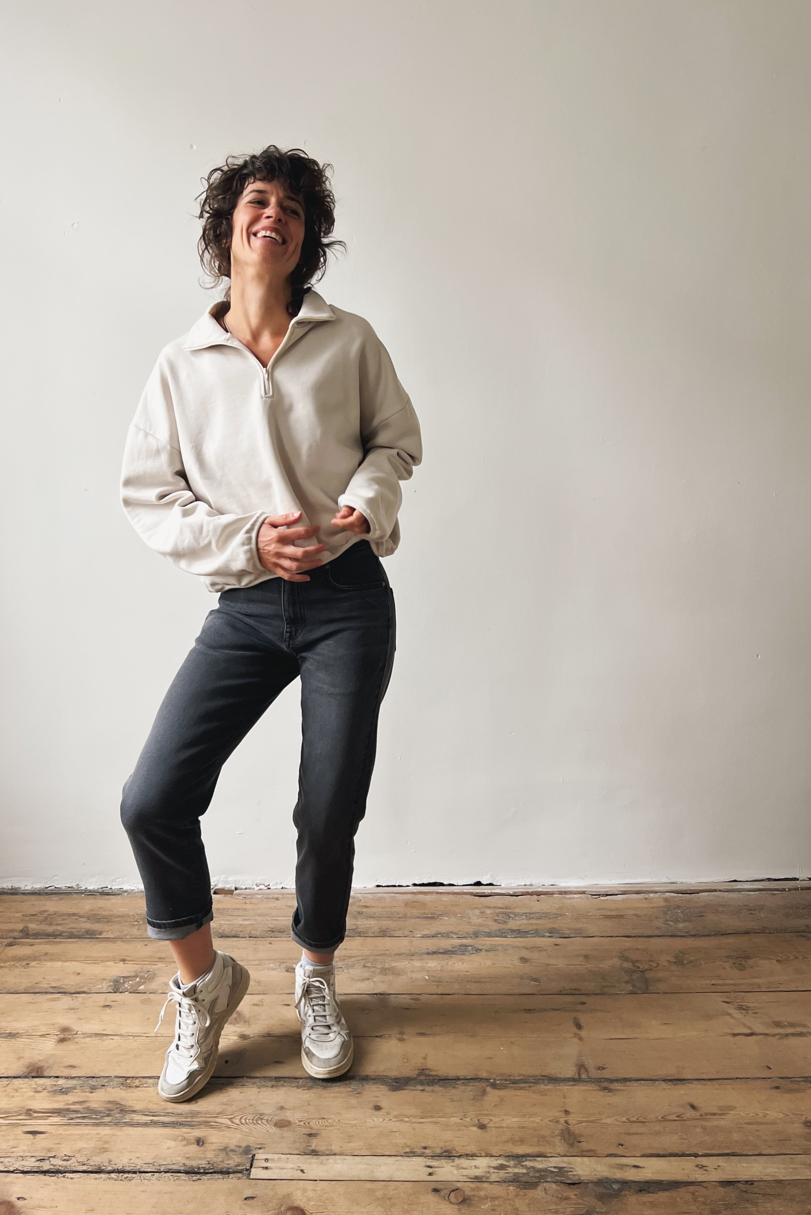 Woman wearing a beige sweater and dark grey boyfriend jeans standing on a wooden floor with a white wall background
