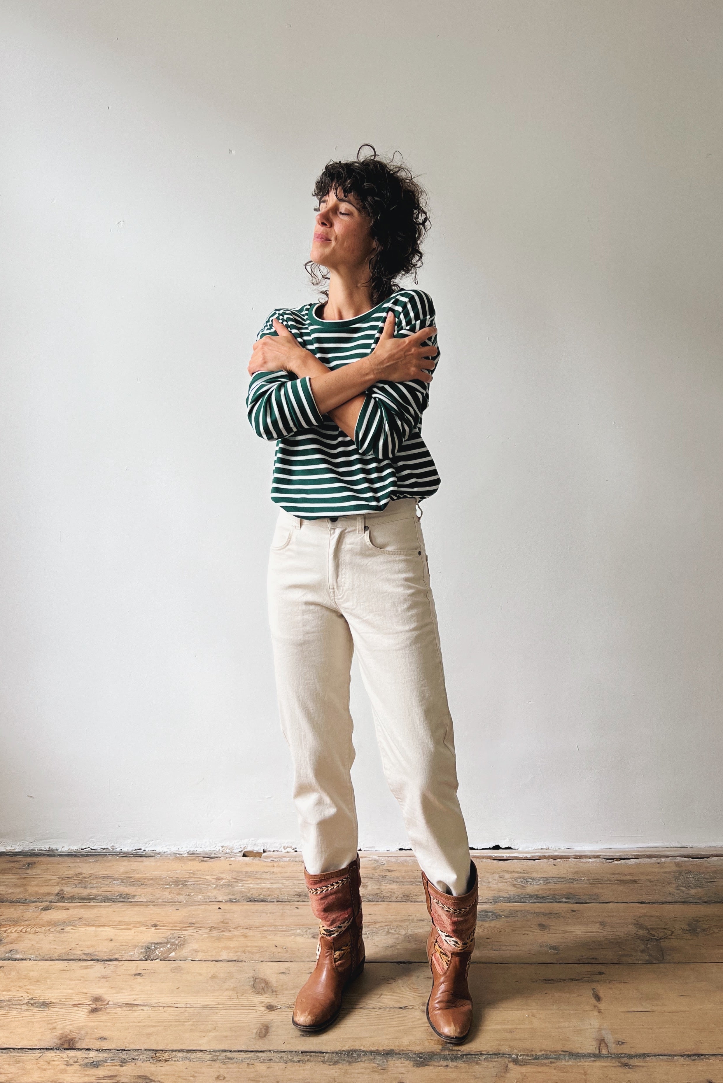 Person wearing a striped shirt, oat jeans, and brown boots standing against a plain wall.